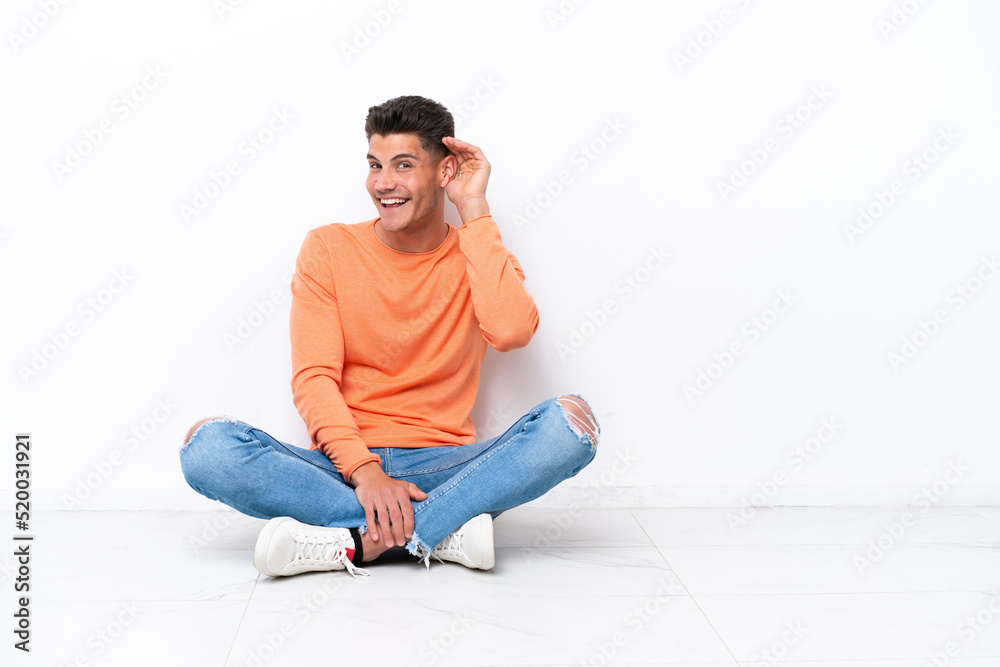 Young man sitting on the floor isolated on white background listening to something by putting hand on the ear Young man sitting on the floor isolated on white background listening to something by putting hand on the ear