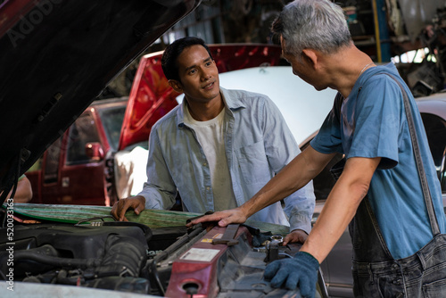 senior Asian mechanic looks for parts to fix a broken engine and stands in front of the car feeling tired talking wiht man about engine broken in the garage.