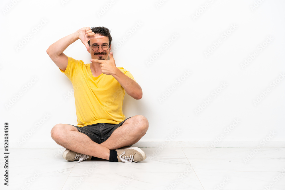 Fototapeta premium Young caucasian man sitting on the floor isolated on white background focusing face. Framing symbol