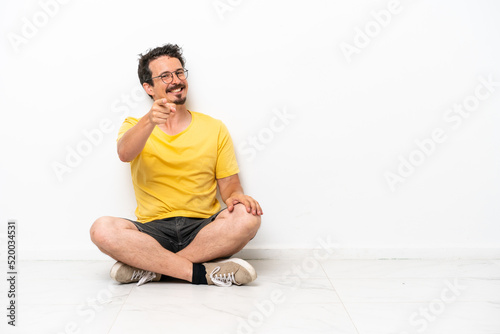 Young caucasian man sitting on the floor isolated on white background points finger at you with a confident expression