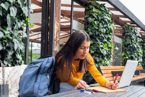 Young female student writing in her notebook next to her laptop sitting at a table outside
