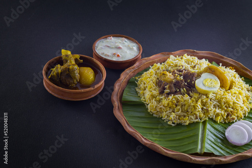 Kolkata style mutton biryani with potato and egg served on clay plate and banana leaf with mutton curry and curd raita. shot against black background.