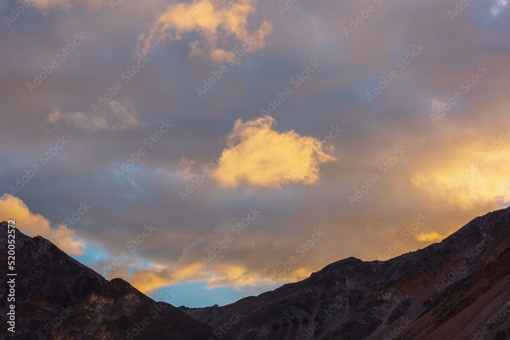 Scenic landscape with dark mountain range silhouette under evening blue sky with vivid orange sunset clouds. Sunlit orange cirrus clouds in sunset sky above mountains silhouettes at changeable weather