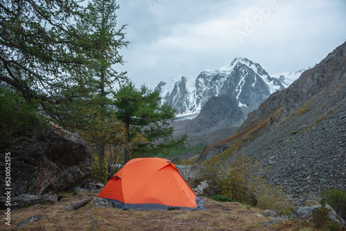 Dramatic landscape with alone orange tent on forest hill among rocks and autumn flora with view to large snowy mountain range under cloudy sky. Lonely tent and fading autumn colors in high mountains.