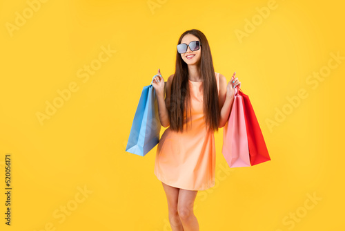 Fashion woman wearing summer dress and sunglasses holding shopping bags on a yellow background with copy space. Holidays, summer sales, discounts, shopping concept.