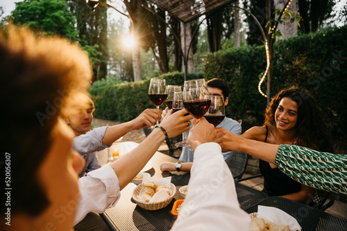 Group of friends outside eat and have fun together toasting with red wine glasses at sunset at the hotel restaurant where are staying - Cheerful people laugh and joke evening  - Party at home