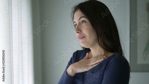 Anxious young woman trying to calm herself standing by window looking out. Person biting nail touching chest with hand