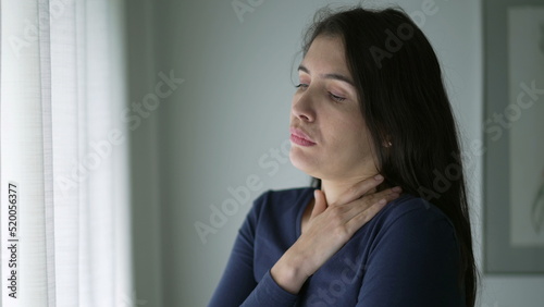 Anxious young woman trying to calm herself standing by window looking out. Person biting nail touching chest with hand © Marco