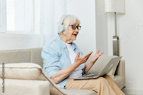 an old woman, carried away by a conversation, is sitting on a cozy sofa and actively communicating via video with headphones on her head while looking at a laptop monitor © Tatiana