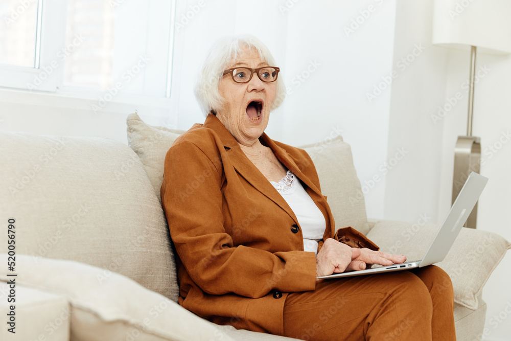 a shocked elderly lady is sitting on a sofa in a bright apartment and holding a laptop on her lap while sitting in a brown stylish suit with her mouth wide open in surprise a shocked elderly lady is sitting on a sofa in a bright apartment and holding a laptop on her lap while sitting in a brown stylish suit with her mouth wide open in surprise