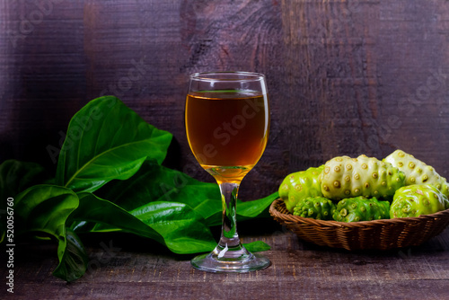 Noni juice in wineglass and noni fruit on basket with green leaf on rustic wooden background. 