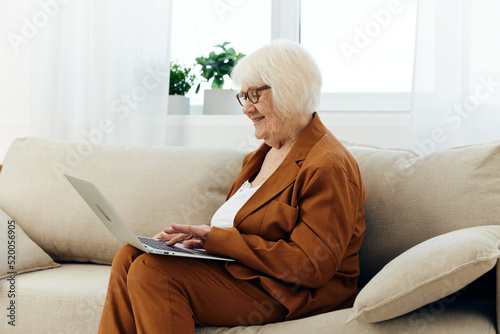 a nice, sweet elderly woman is sitting on the sofa in a bright apartment typing text in a laptop holding it on her lap and pleasantly smiling with happiness © Tatiana
