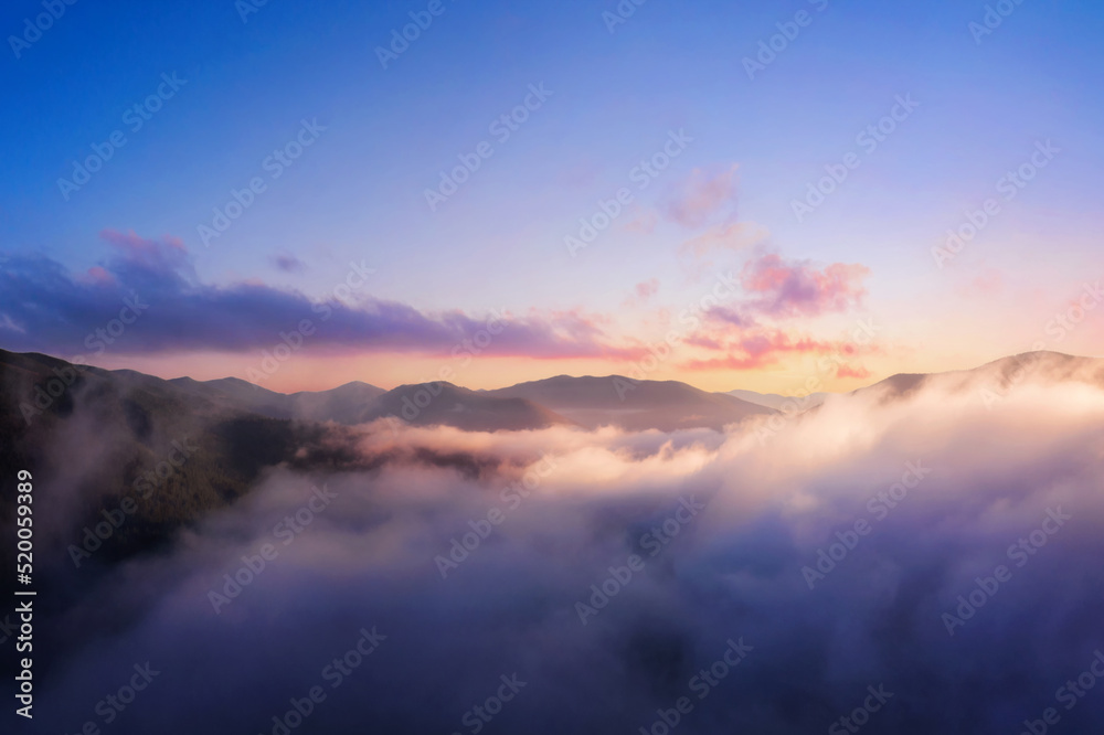 Obraz premium Mountains in low clouds. Mountain landscape with dramatic cloudy sky, peaks in fog and bright sunlight at dawn in the morning. Aerial view. Travel and tourism.