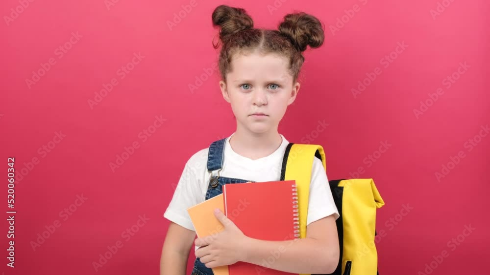 Dislike concept. Dissatisfied little kid girl holding two notebooks gesturing thumbs down disapproving something, wearing yellow backpack, posing isolated over red color background wall in studio