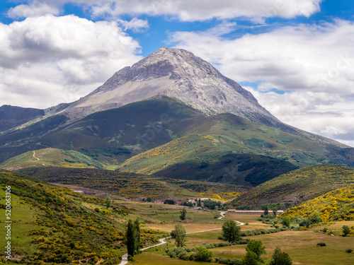 Montaña rocosa con forma piramidal bajo un cielo azul de nubes blancas y verdes prados con un camino que nos conduce a un pueblo
