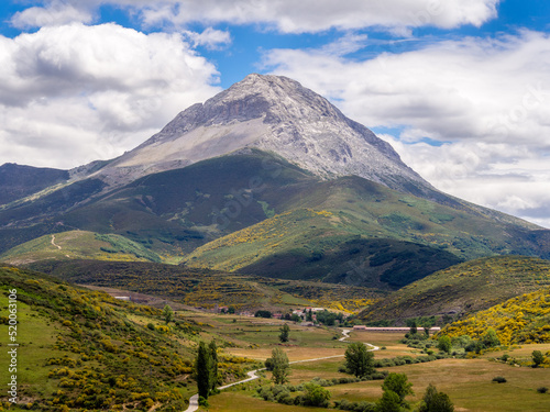 Montaña rocosa con forma piramidal bajo un cielo azul de nubes blancas y verdes prados con un camino que nos conduce a un pueblo
 photo