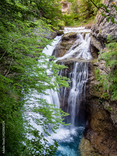 Cascada de alta montaña cae entre paredes verticales y vegetación de ribera con hojas verdes.