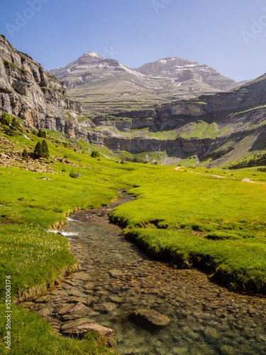 Paisaje alpino donde un riachuelo recorre un valle verde dirección a dos montañas bajo un cielo azul.