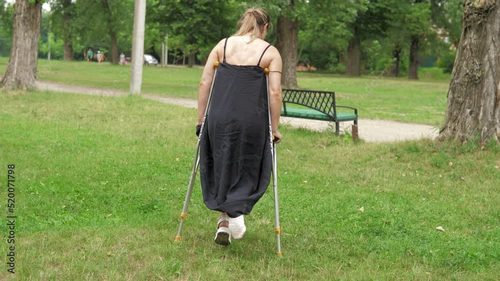 A young girl with a cast walks around the park on crutches on the lawn
