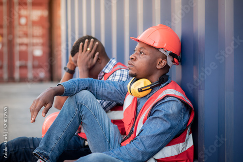 2 African American black man worker wearing safety uniform workwear looking hopeless and stressed, sitting beside a shipping container, unemployment due to the economic recession No cargo activities. © pornchai