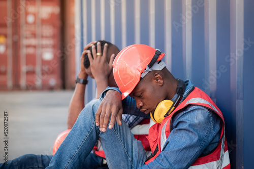 Close-up 2 African American black man worker wearing safety uniform workwear and sitting on the floor leaning against the container because there is no work to do, economic recession due to inflation. © pornchai