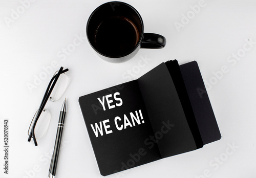 YES WE CAN written text in small black notebook with coffee , pen and glasess on white background. Black-white style