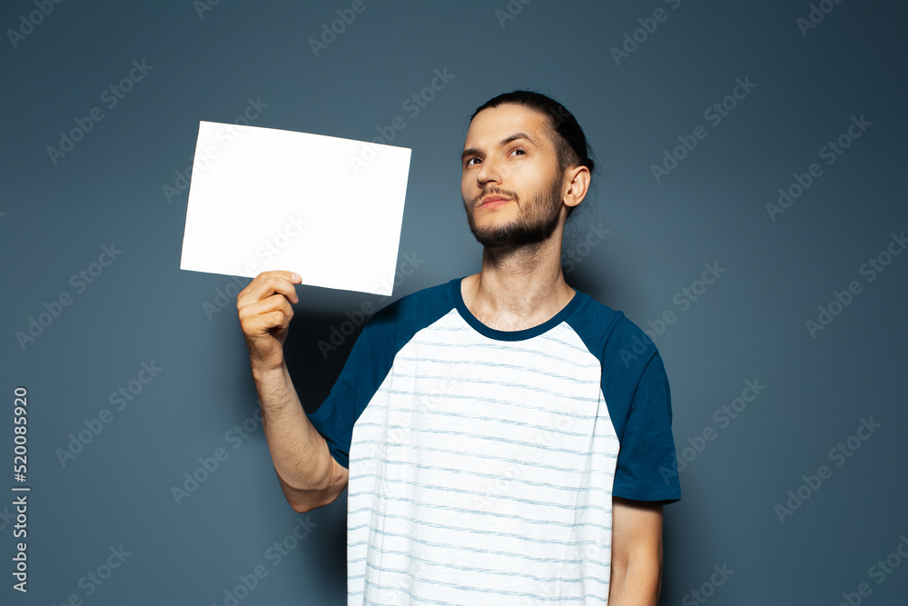 Studio portrait of thoughtful young man holding white empty paper board on blue color. Looking up. Studio portrait of thoughtful young man holding white empty paper board on blue color. Looking up.