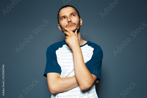 Studio portrait of young thoughtful man looking up with hand under chin on blue background.
