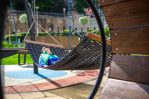 Cute little blond caucasian boy relaxing and having fun in multicolored hammock in backyard or outdoor playground. Summer active leisure for kids. Child swinging on hammock. Activities for children