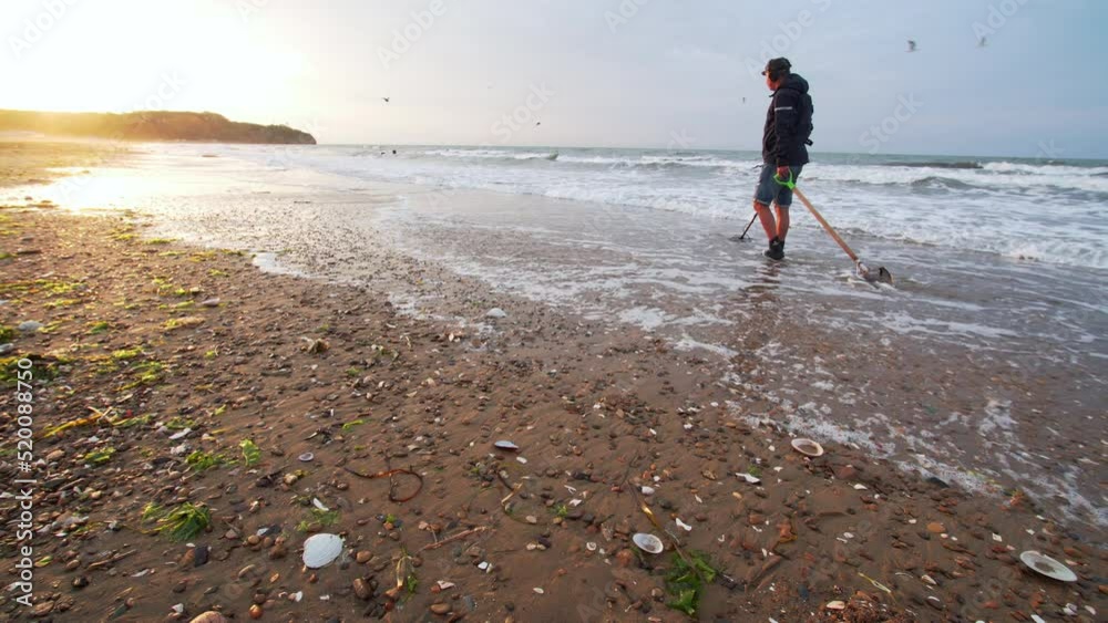 A man is looking for treasure with electronic metal detector on the sea beach in the morning. He is digging the sand and sieving it looking for the lost golden goods