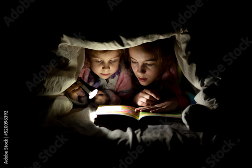two sisters are reading a book under the covers at night with a flashlight