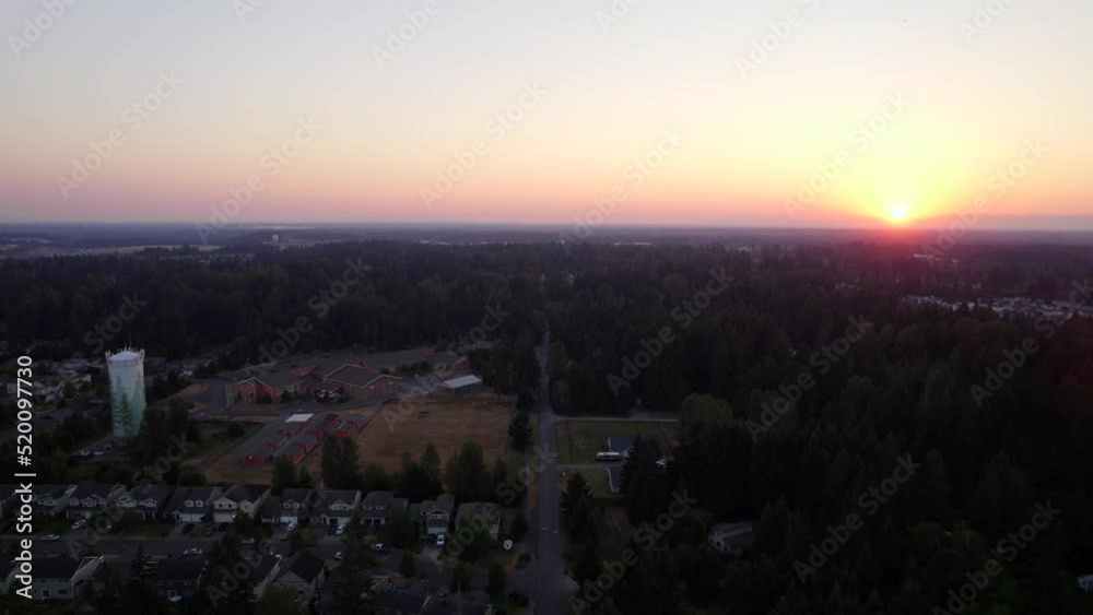 Flying backwards over a street in a residential neighborhood of houses, a water tower, and a school as the sun begins to set over the city of Puyallup, Washington.