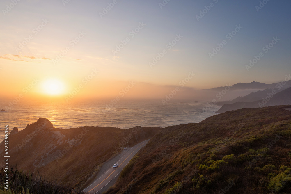 Golden sunset over California State Route 1, on the Pacific Ocean. Pacific Coast, Central California near Gorda. Car traveling south on Highway One as the sunset glows warm tones on the hillside