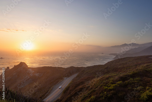 Golden sunset over California State Route 1, on the Pacific Ocean. Pacific Coast, Central California near Gorda. Car traveling south on Highway One as the sunset glows warm tones on the hillside