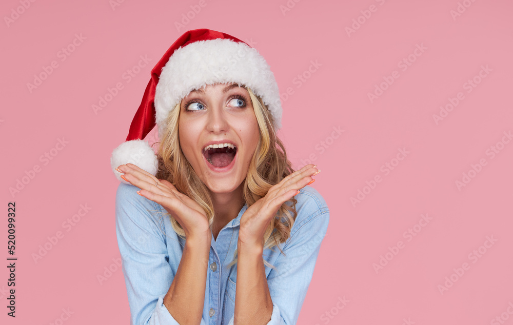 Portrait of a cheerful joyful girl in casual clothes, Santa hat on a pink background looks to the right. Wow face. Concept of Christmas and New year holidays Portrait of a cheerful joyful girl in casual clothes, Santa hat on a pink background looks to the right. Wow face. Concept of Christmas and New year holidays