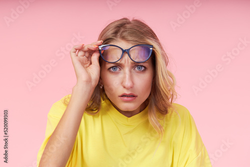 Portrait of young excited woman in glasses  yellow casual t-shirt on pink background. Surprised and incredulous looks wide open mouth and eyes