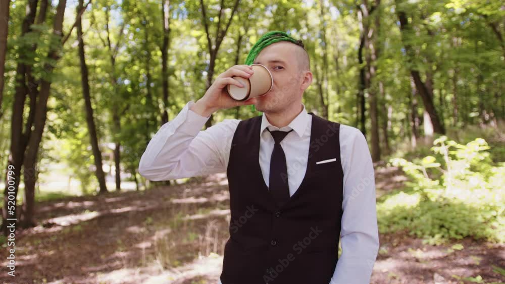Young man with dreadlocks drinking coffee in forest park. Stylish hipster in business suit with green hairstyle enjoying hot drink from paper cup in summertime.