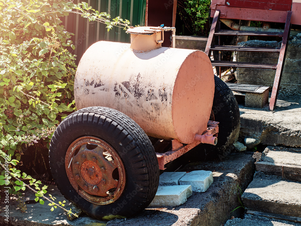 Water tank trailer on wheels. Barrel on wheels. A light pink rusting iron tank on wheels stands in the countryside on concrete slabs. Water tank. Water tank trailer on wheels. Barrel on wheels. A light pink rusting iron tank on wheels stands in the countryside on concrete slabs. Water tank.