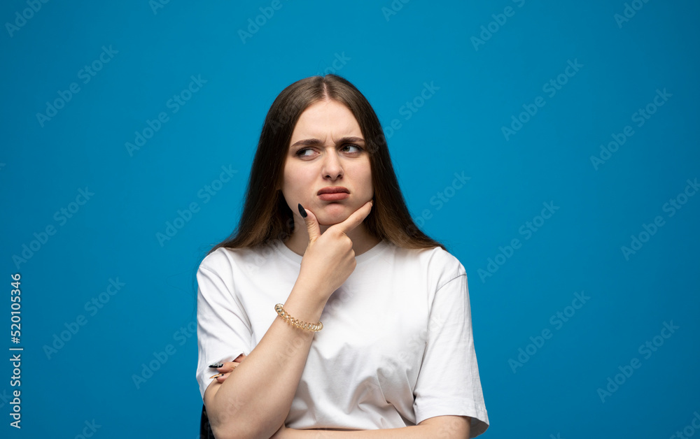 Portrait of upset young beautifil brunette girl with long hair in a white t-shirt standing with puffy cheeks isolated over blue background. Emotion of angry and upset. Portrait of upset young beautifil brunette girl with long hair in a white t-shirt standing with puffy cheeks isolated over blue background. Emotion of angry and upset.