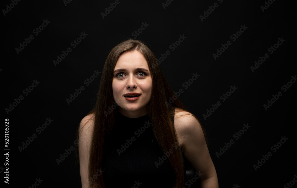 Fototapeta premium Portrait of upset young beautifil brunette girl with long hair in a black t-shirt in a black room. Emotion of angry and upset.