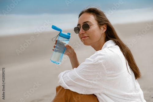 Beautiful woman in sunglasses sitting near sea, drinking water and relax during vacation