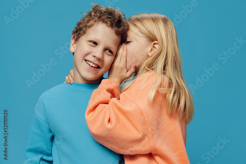  children of school age in colored sweaters stand on a blue background and the girl tells the boy a secret in the ear while standing sideways to the camera. The theme of friendship between children