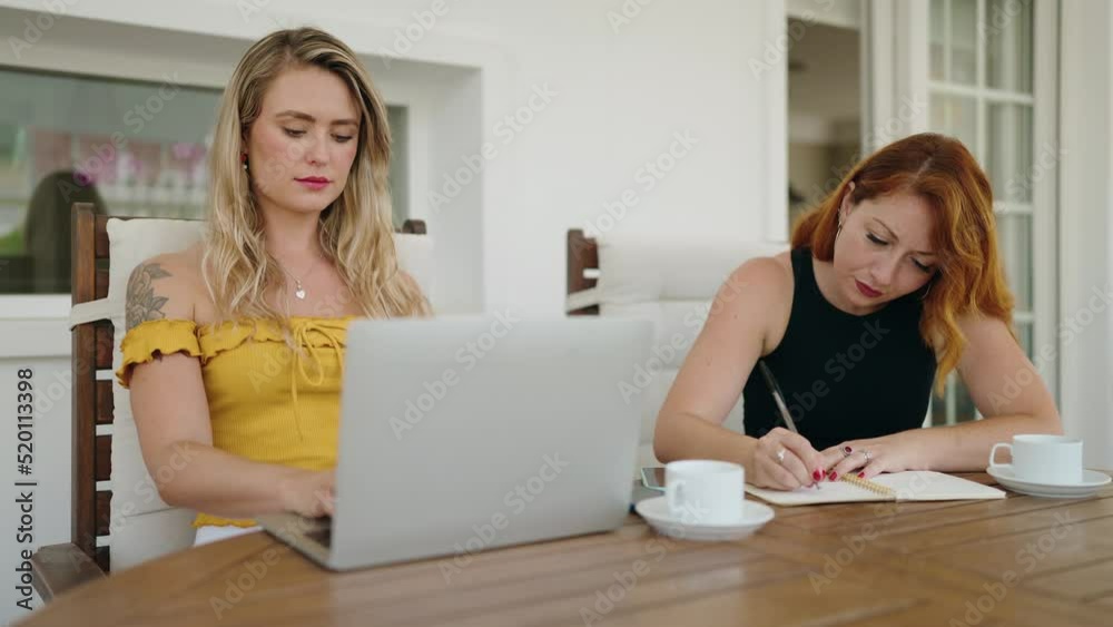 Two women using laptop writing on notebook sitting on table at home terrace Two women using laptop writing on notebook sitting on table at home terrace