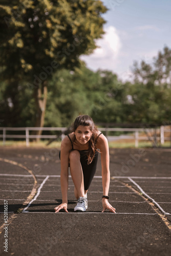 Concentrated Caucasian girl athlete in dressed in sportswear: shorts and top, sneakers with fitness tracker preparing for run on track at stadium in summer. Happy female outdoor sport concept