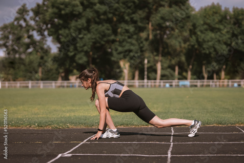 Concentrated Caucasian girl athlete in dressed in sportswear: shorts and top, sneakers with fitness tracker preparing for run on track at stadium in summer. Happy female outdoor sport concept