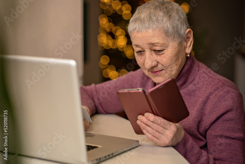 An elderly woman sitting in front of a laptop holds a bank card in her hands and makes online purchases.