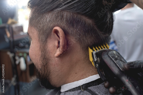 Canvas Print A barber with black gloves uses a white hair clipper with a yellow comb guide to trim the side scalp of a customer