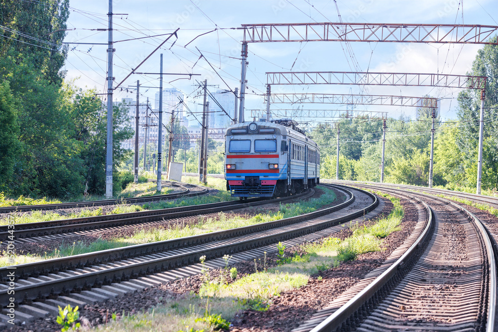 Naklejka premium The outgoing old train rides along the railroad against the backdrop of city buildings in a sunny haze.
