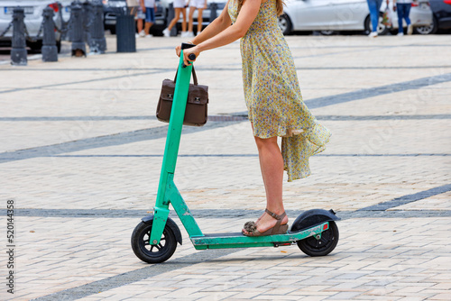 A girl in a light dress rides an electric scooter along a cobbled city square. The concept of eco-friendly free movement.