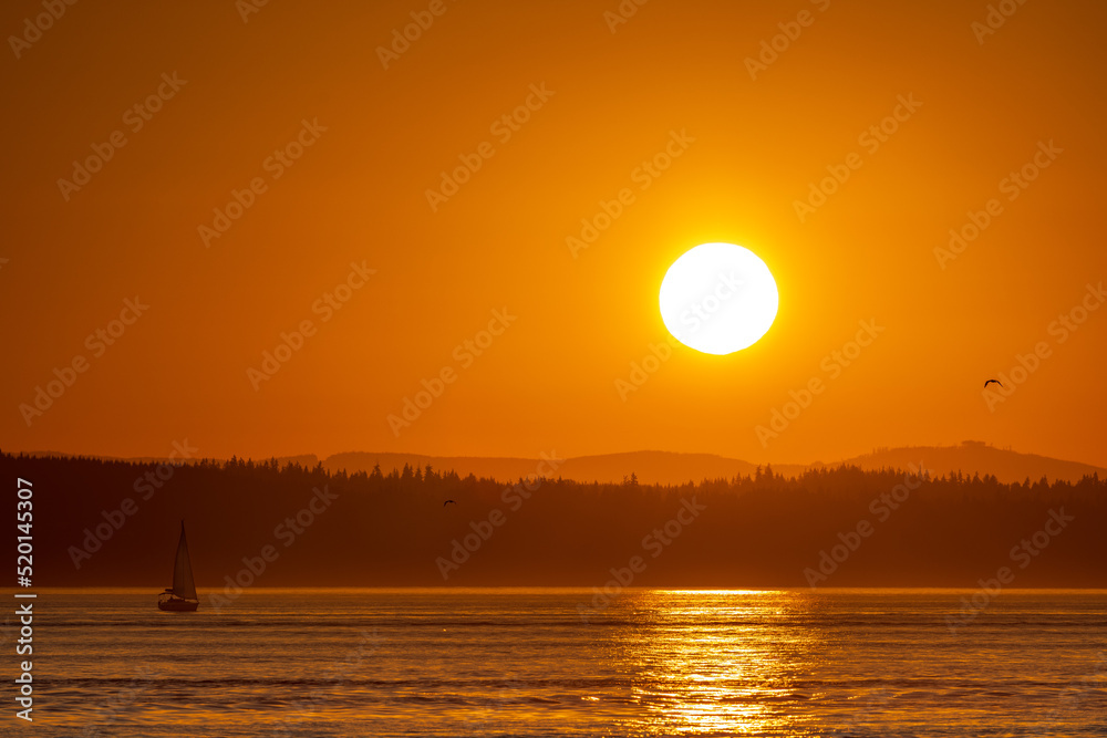 A brilliant orange sun sets over Puget Sound with silhouetted hills in the background and reflections on the ocean in the foreground